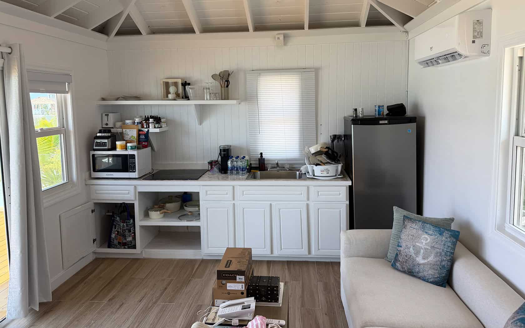 Bright white cabin kitchenette with microwave, coffee maker, sink, and a tall stainless fridge on the right; open shelving above and a sofa with a blue anchor pillow to the side.