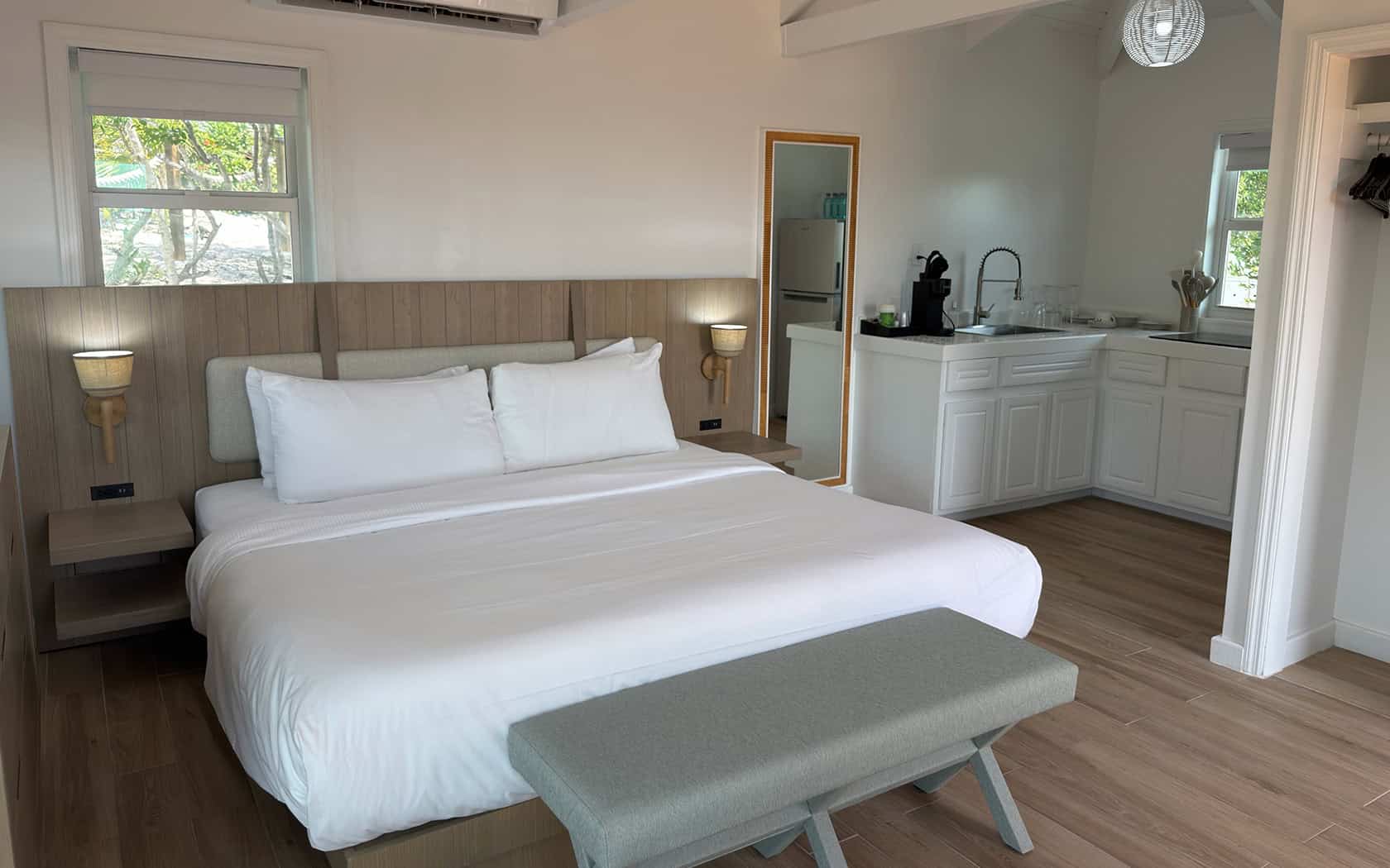 Queen bed in a bright room with white linens and a light wood headboard; bench at the foot and kitchenette visible in the background.