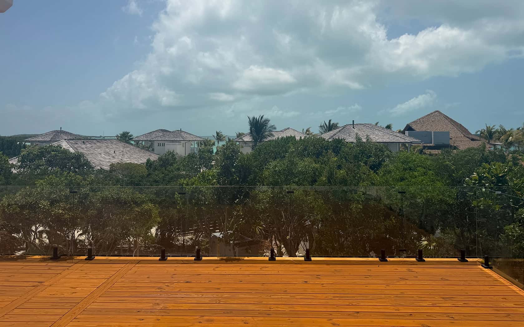 Wooden deck with glass railing overlooking a tropical neighborhood with palm trees and thatched-roof houses under a blue sky