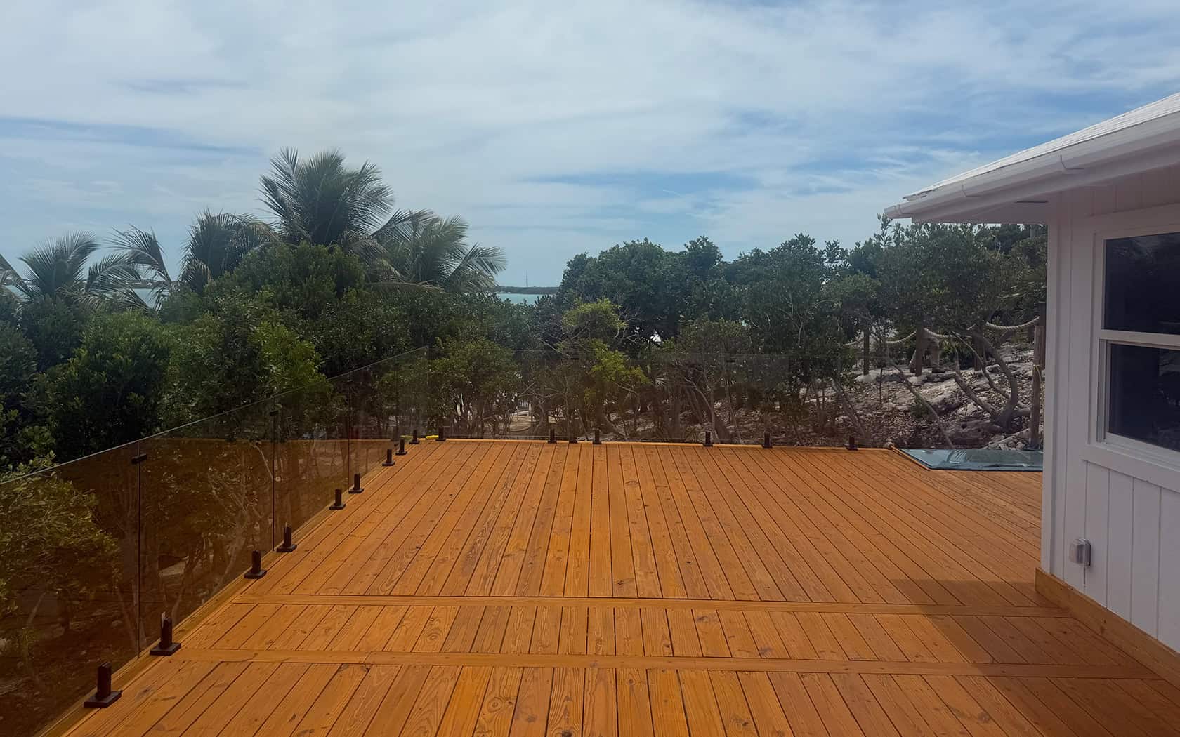 Wooden deck with glass railing overlooking tropical trees and distant ocean, with a white house on the right.