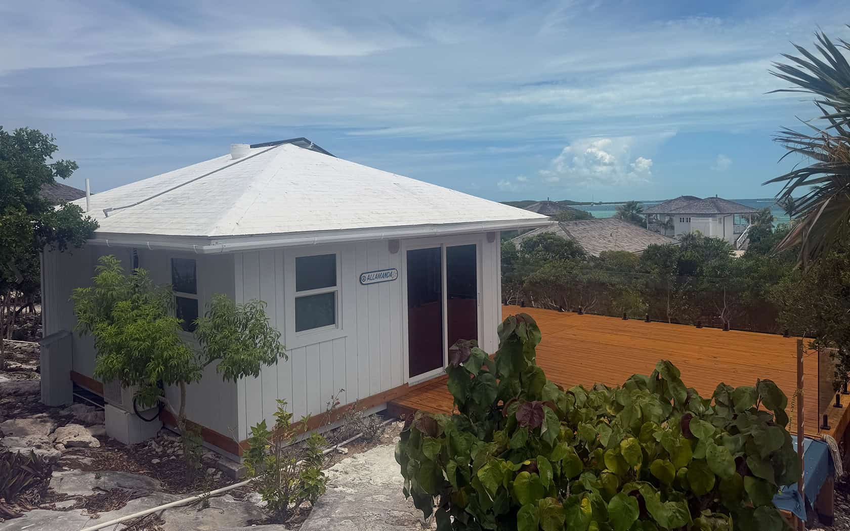 White beach bungalow with a pale pyramid-like roof, orange wooden fence, and tropical plants; ocean and neighboring houses in the background.