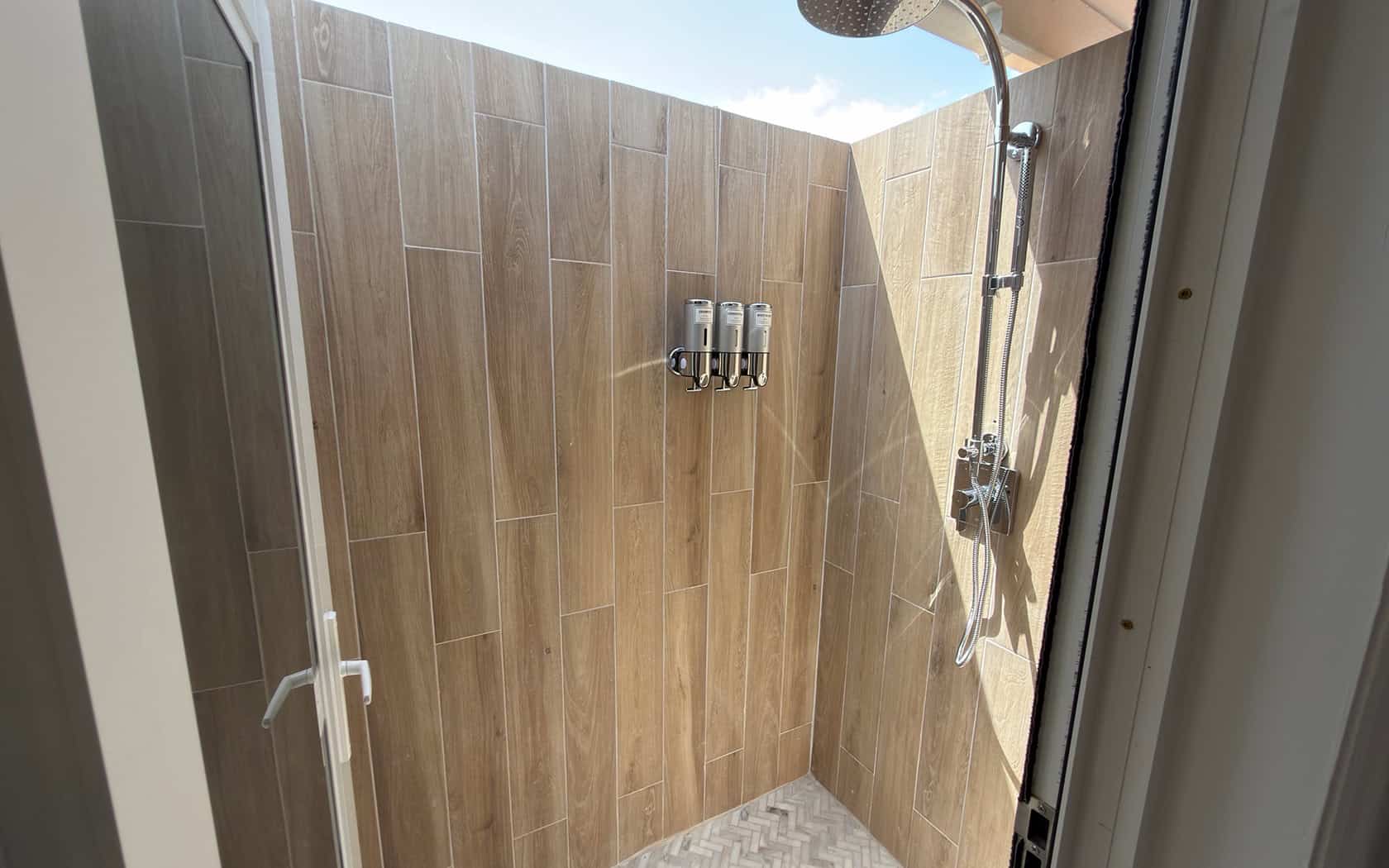 Outdoor shower area with wood-toned tiles, wall-mounted three‑bottle dispensers, and a ceiling rainfall shower head