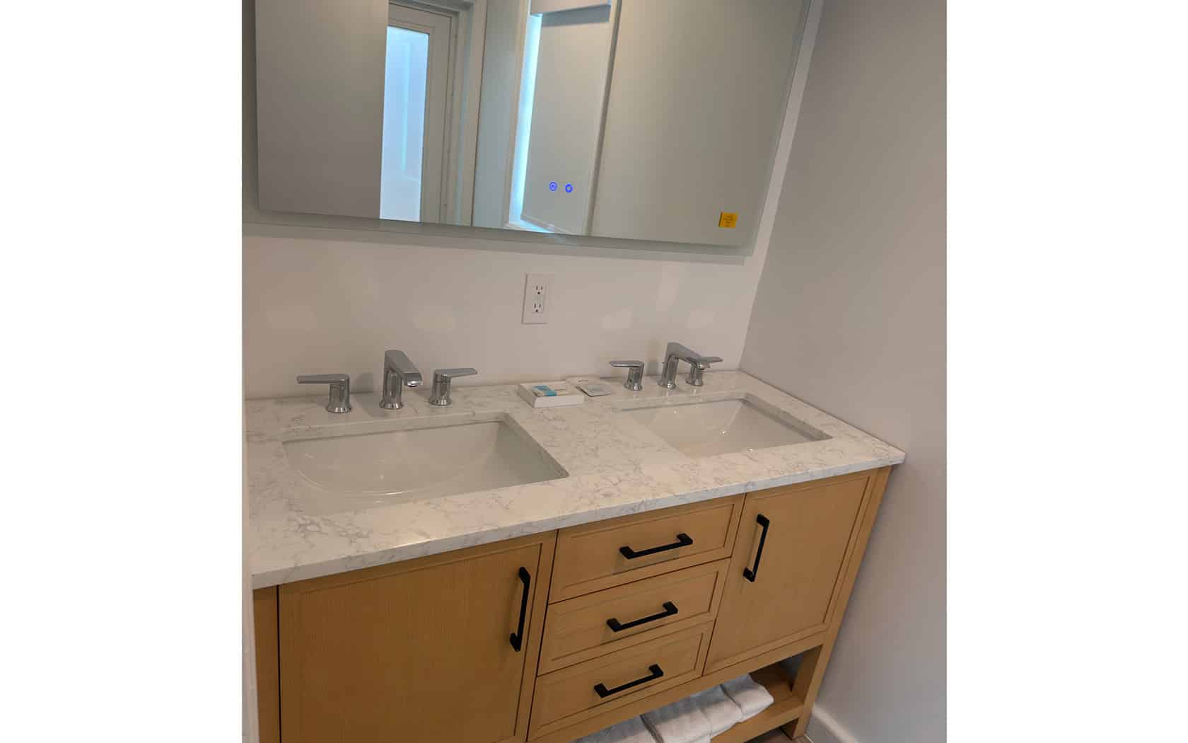 Double-sink bathroom vanity with marble countertop, chrome faucets, and light wood cabinets beneath. A large mirror runs above the counter.